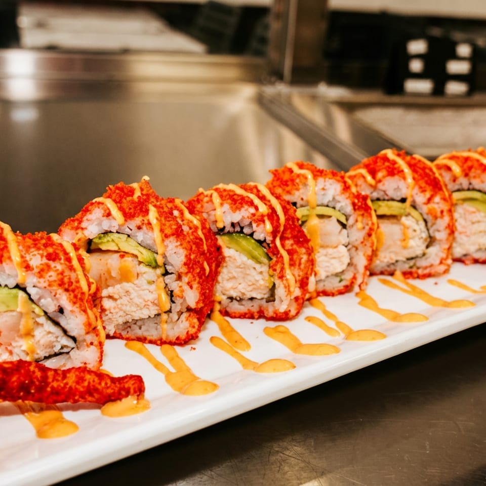 Sushi roll slices topped with orange sauce and red roe, served on a rectangular white plate with decorative sauces.