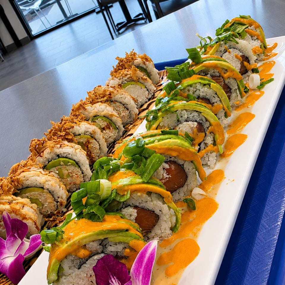 Long platter of sushi rolls topped with avocado, scallions, crispy garnish, orange sauce, and purple orchids in a bright restaurant.