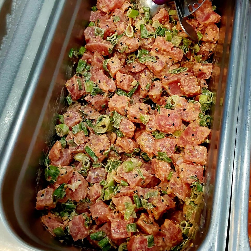 Tray of diced seasoned sausage mixed with green onions and black sesame seeds, served with purple tongs.