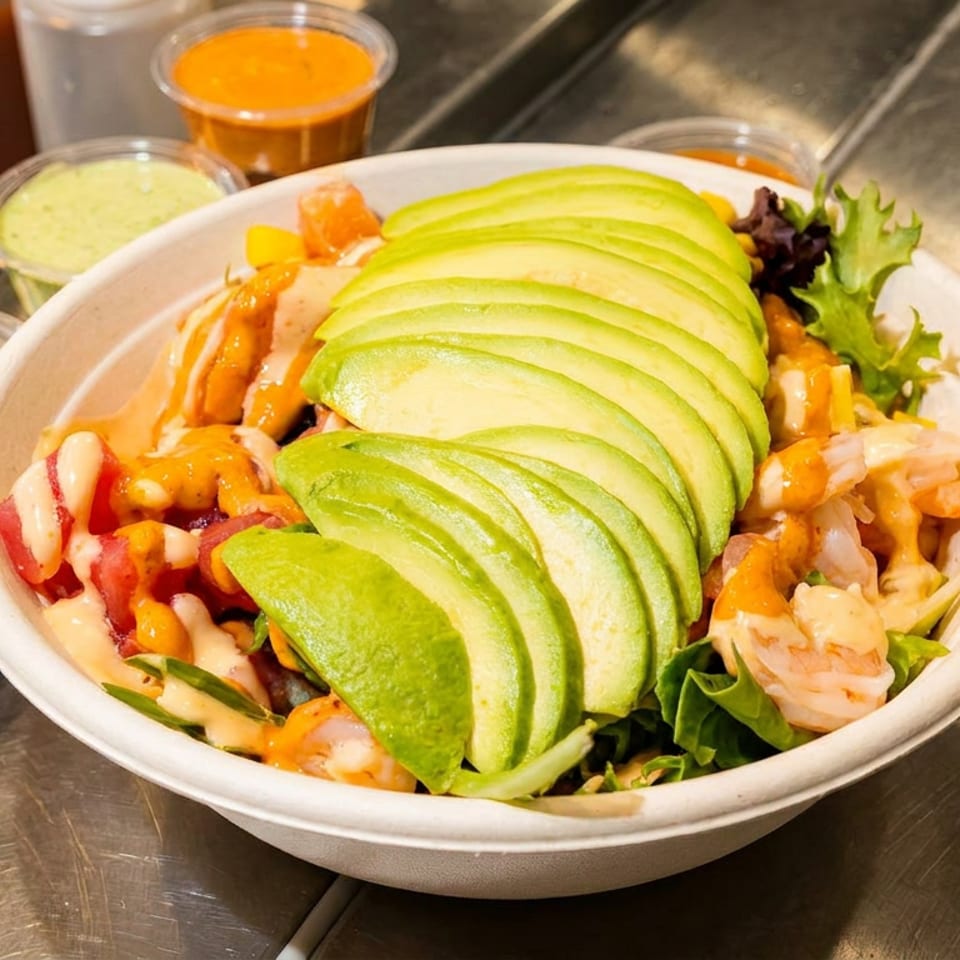 Bowl of greens topped with sliced avocado, shrimp, vegetables, and creamy orange dressing at a restaurant preparation counter.
